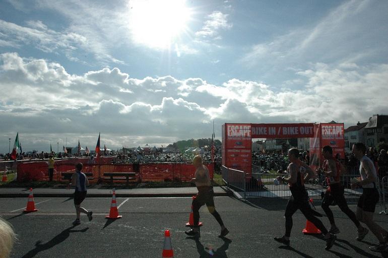 Triathletes running toward a red bike transition station under sunny skies with crowds in background.
