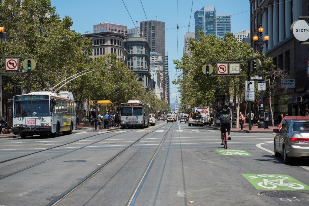 Market Street at Sixth Street looking east with cable car tracks, transit bus, cyclists, and downtown San Francisco skyline.