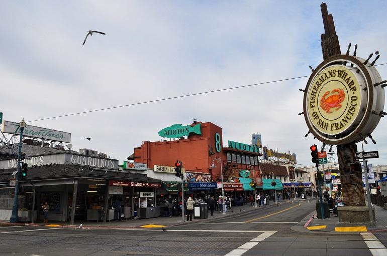 Fisherman's Wharf street scene with historic ship wheel sign, storefronts, and pedestrians at Jefferson and Taylor streets.