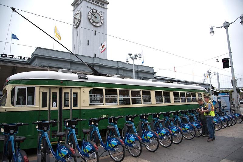 Green and cream Muni streetcar with blue Ford GoBike bicycles lined up in front of the Ferry Building clock tower.