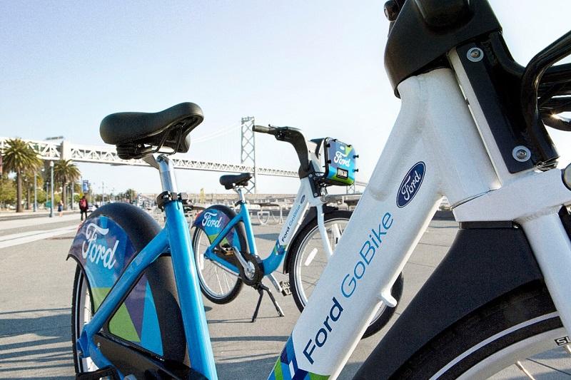 Ford GoBike bicycles docked at a waterfront station with bridge in background.