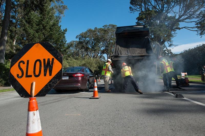 Road construction crew installing speed humps on John F. Kennedy Drive with slow warning sign and traffic control visible.