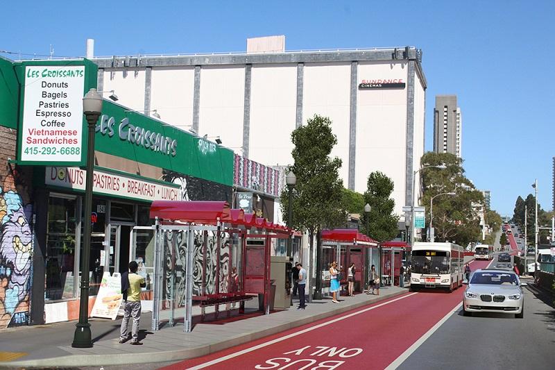 Red transit lane on Geary Boulevard with bus stop, storefronts, and parked vehicles under clear sky.