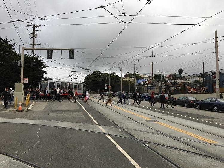 Light rail vehicle stopped at station with pedestrians crossing at intersection under overhead power lines.