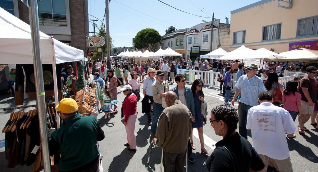 Crowded street festival with white vendor tents, pedestrians, and historic buildings on a sunny day.