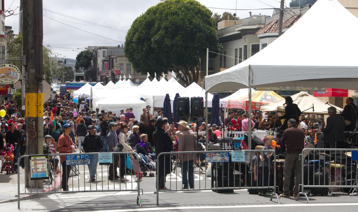 Crowded street festival with white tents, stage, and barriers along San Francisco street lined with trees.