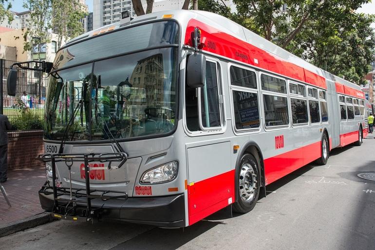 Red and white Muni bus parked on city street next to park with trees and buildings