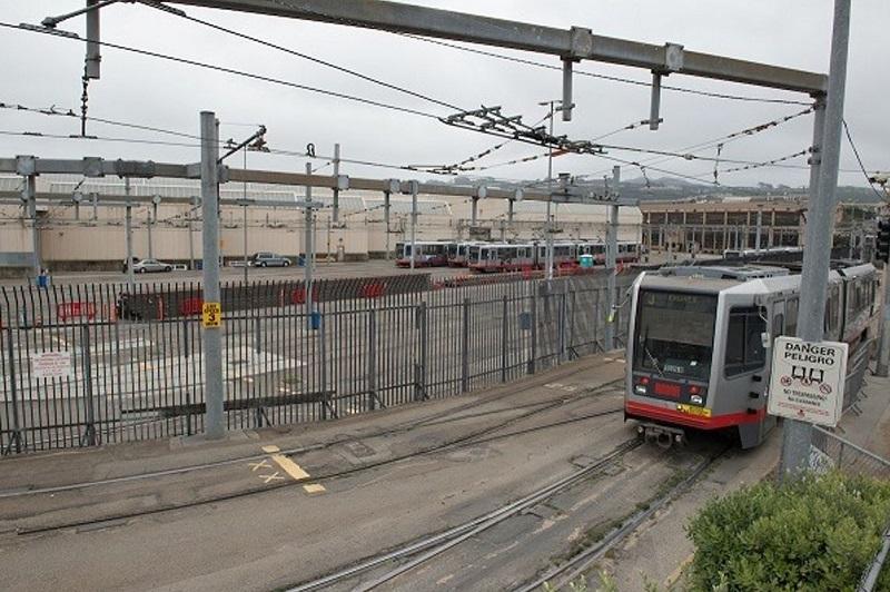Light rail vehicle at Green Division Yard with overhead catenary lines and maintenance facilities visible.