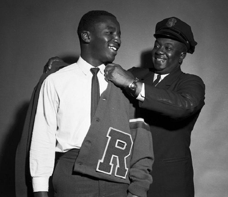 Two men smiling, one adjusting a varsity jacket on the other in a black and white photograph.