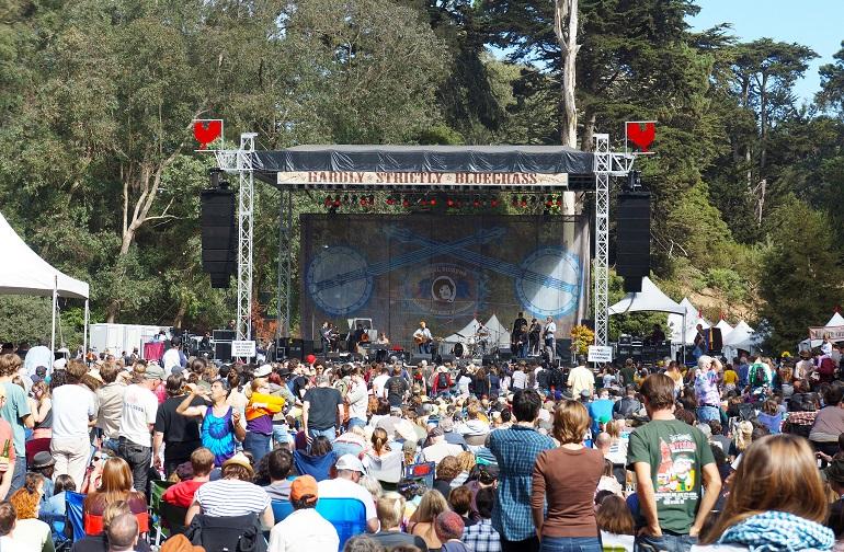 Crowd of people gathered in front of a large outdoor concert stage surrounded by trees and white tents.