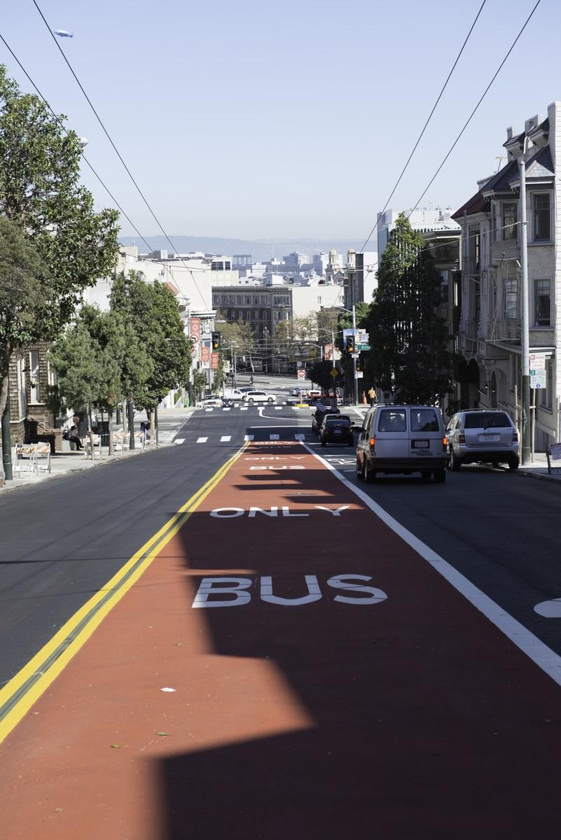 Red transit-only bus lane on Haight Street with yellow border lines and downtown San Francisco skyline in background.