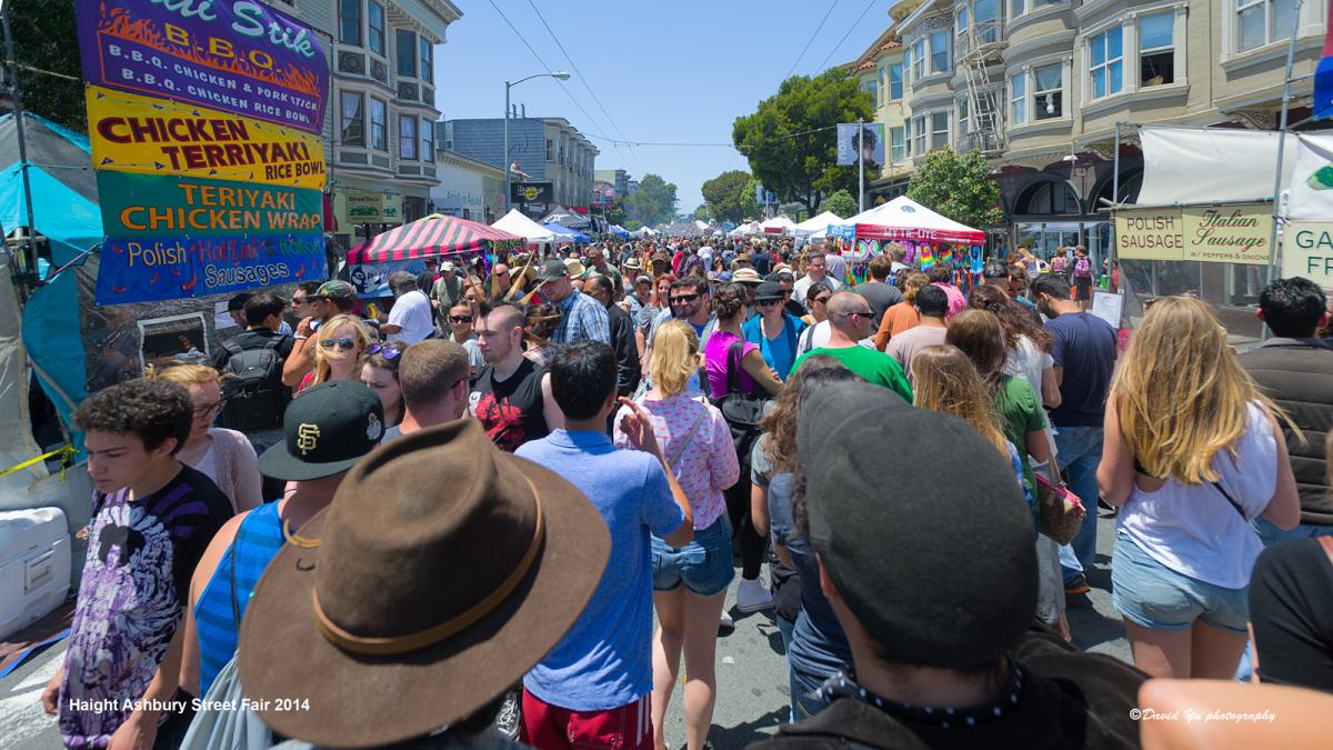 Crowded street fair on Haight Street with food vendors, tents, and hundreds of pedestrians wearing hats and casual clothing.