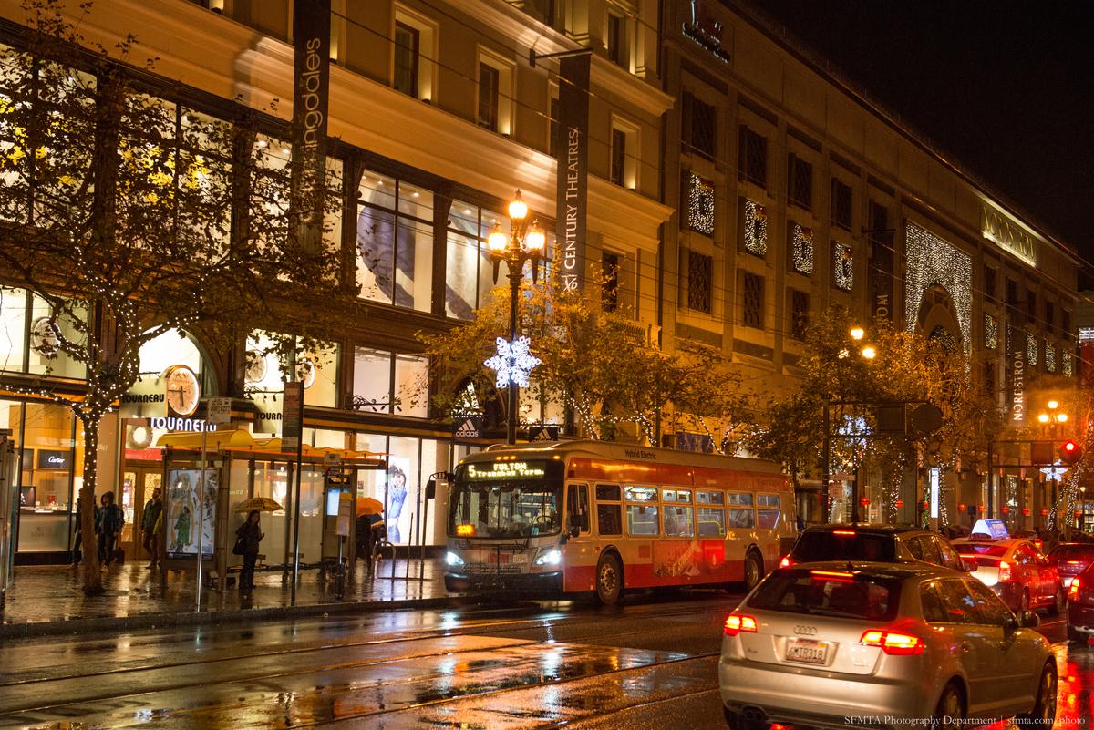 Red bus and cars on wet Market Street at night with illuminated storefronts and holiday lights