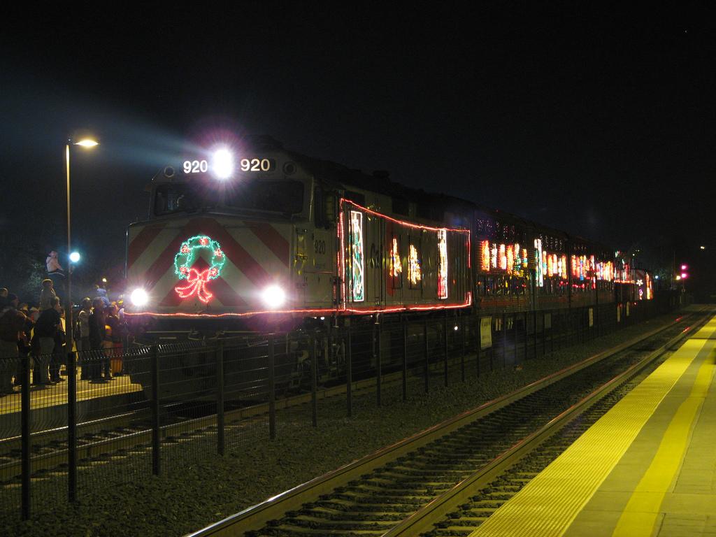 Illuminated holiday train decorated with festive lights and wreath stopped at station platform at night with spectators watching.