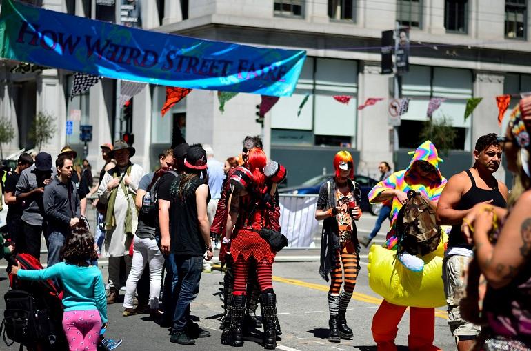 Costumed participants parade on a city street during How Weird Street Faire, with colorful banners and decorations overhead.