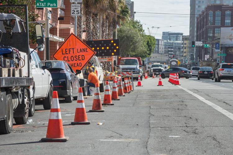 Construction barricades and orange warning sign reading "Left Lane Closed Ahead" on city street with traffic and vehicles.