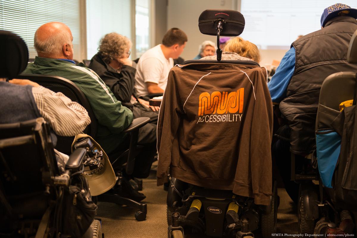 Group of people in wheelchairs and standing, gathered indoors with Muni Accessibility jacket visible on center wheelchair.