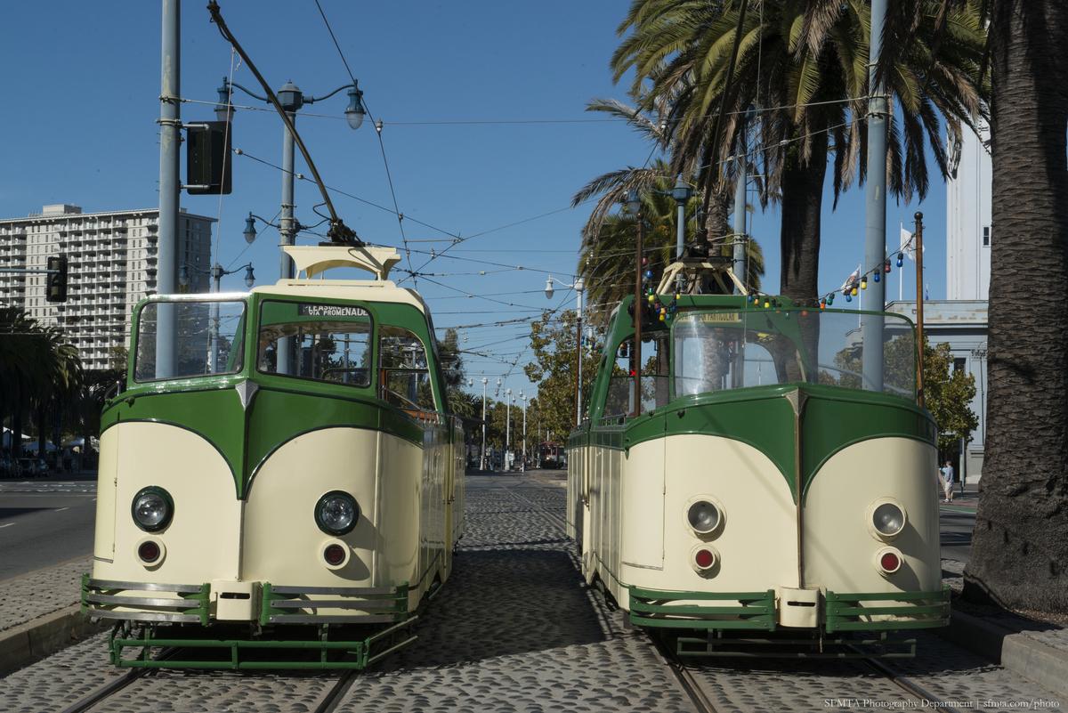 Two cream and green historic streetcars on The Embarcadero with palm trees and buildings in background.