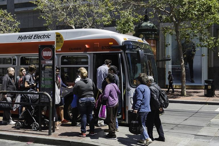 Passengers boarding and exiting a Muni hybrid electric bus on a city street.