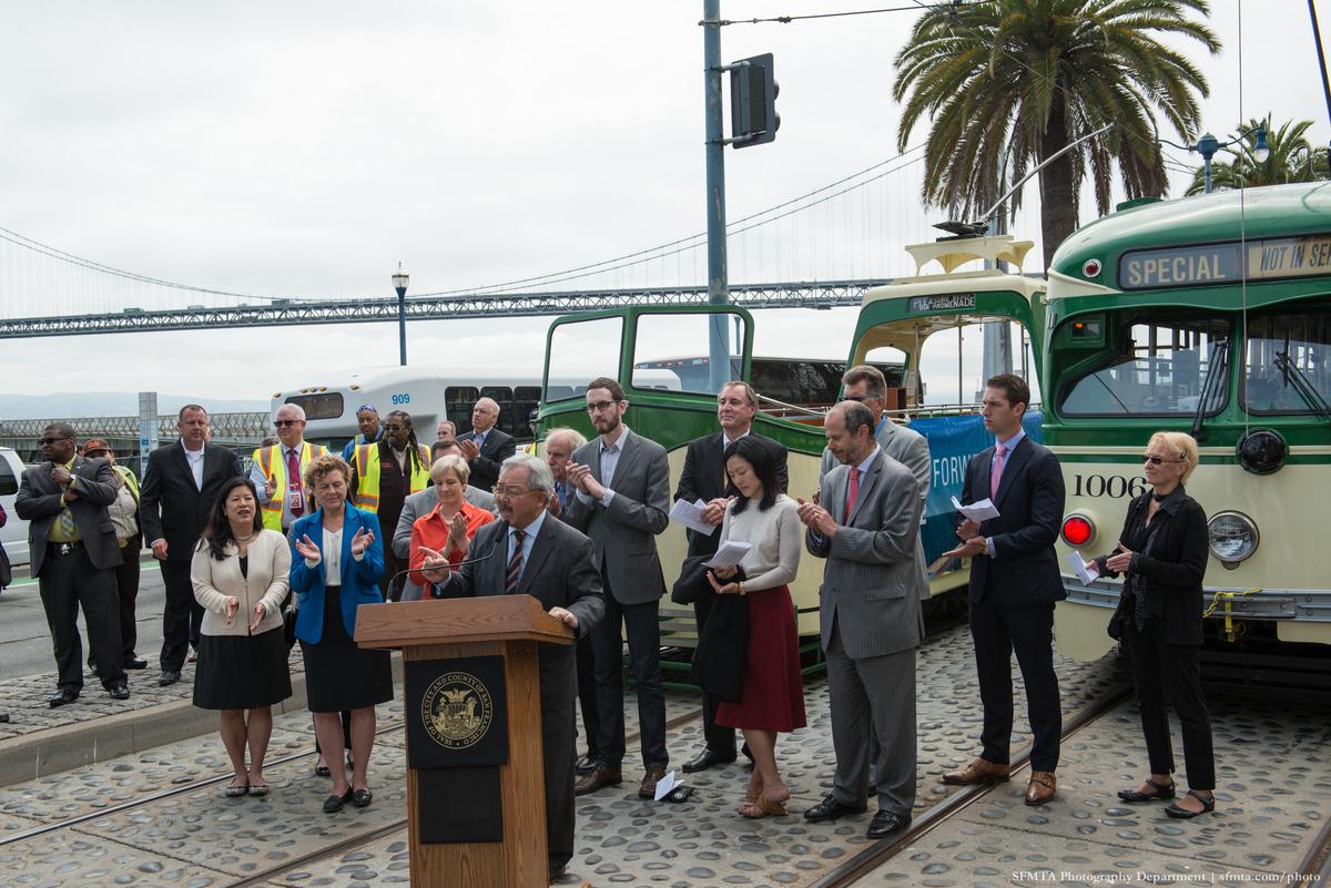 Mayor Lee speaks at podium with dignitaries and historic streetcars on The Embarcadero, Bay Bridge visible behind.