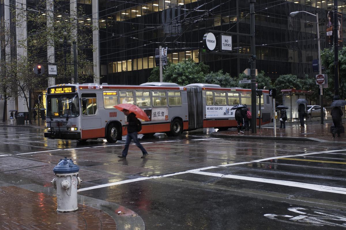 Pedestrian with red umbrella crosses wet downtown street in front of stopped Muni bus during rain.