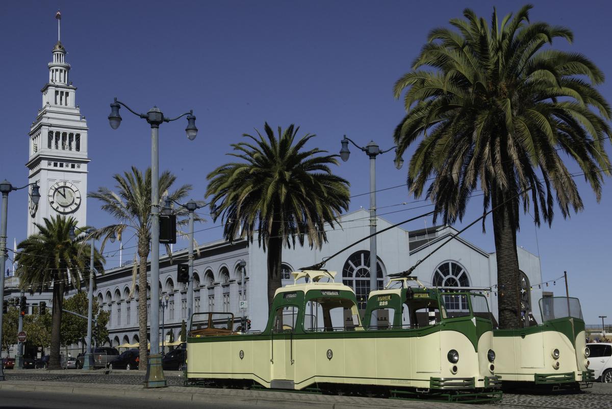Green and cream historic streetcars on The Embarcadero with Ferry Building and palm trees in background.