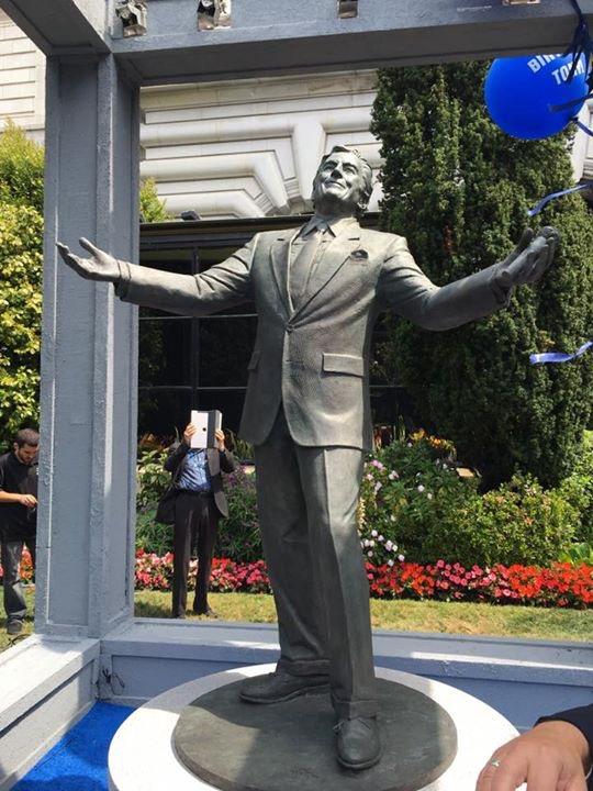 Bronze statue of man with outstretched arms in front of building with gray metal structure.
