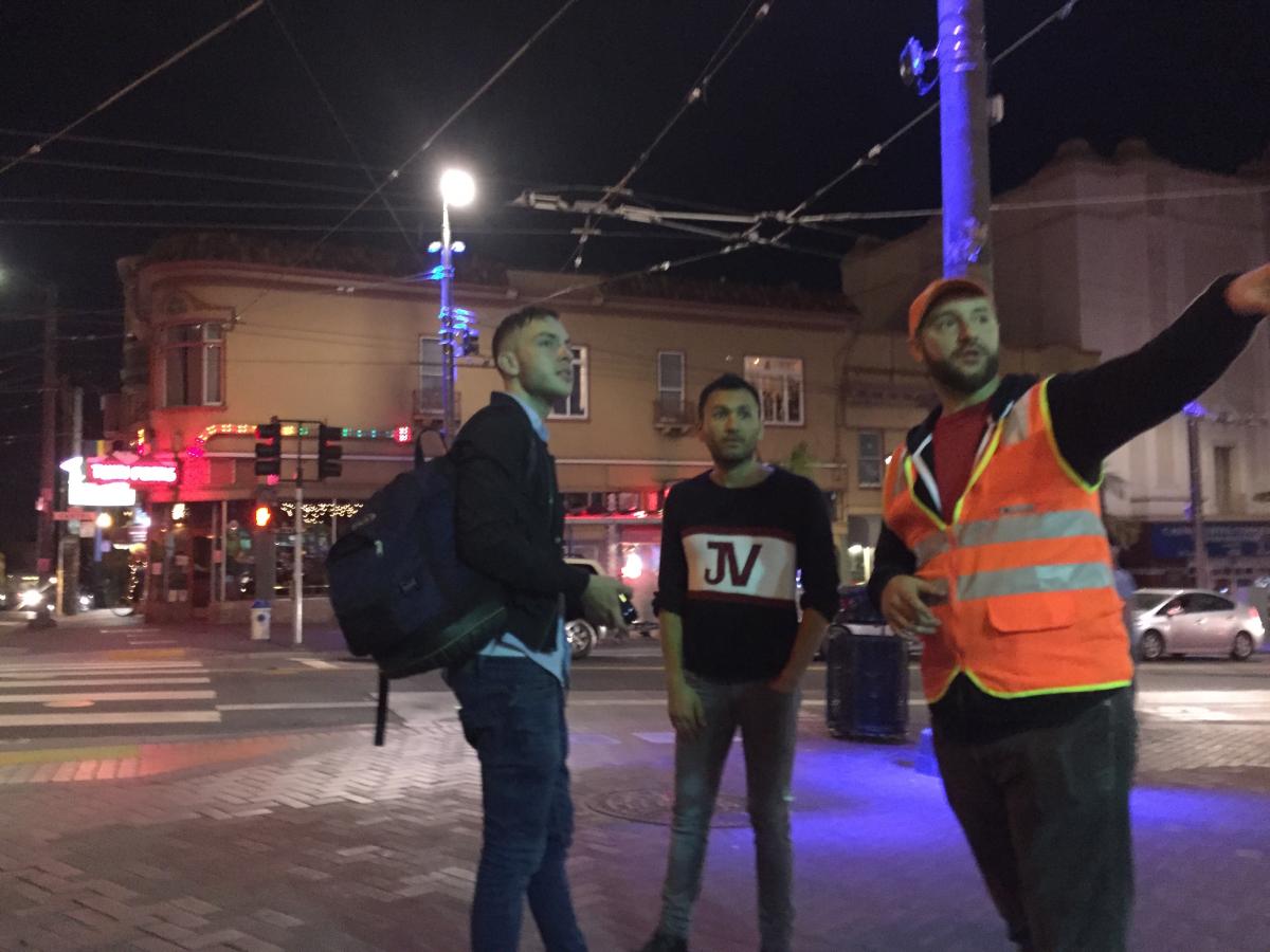 Three men at a nighttime street intersection, one wearing an orange safety vest and hat.