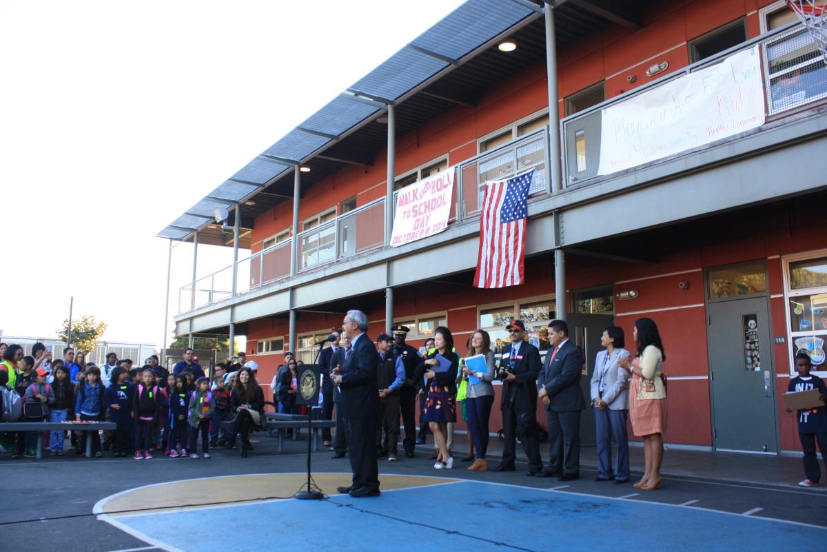 Mayor and city officials stand with students and staff at a school courtyard event, with American flag displayed on building.