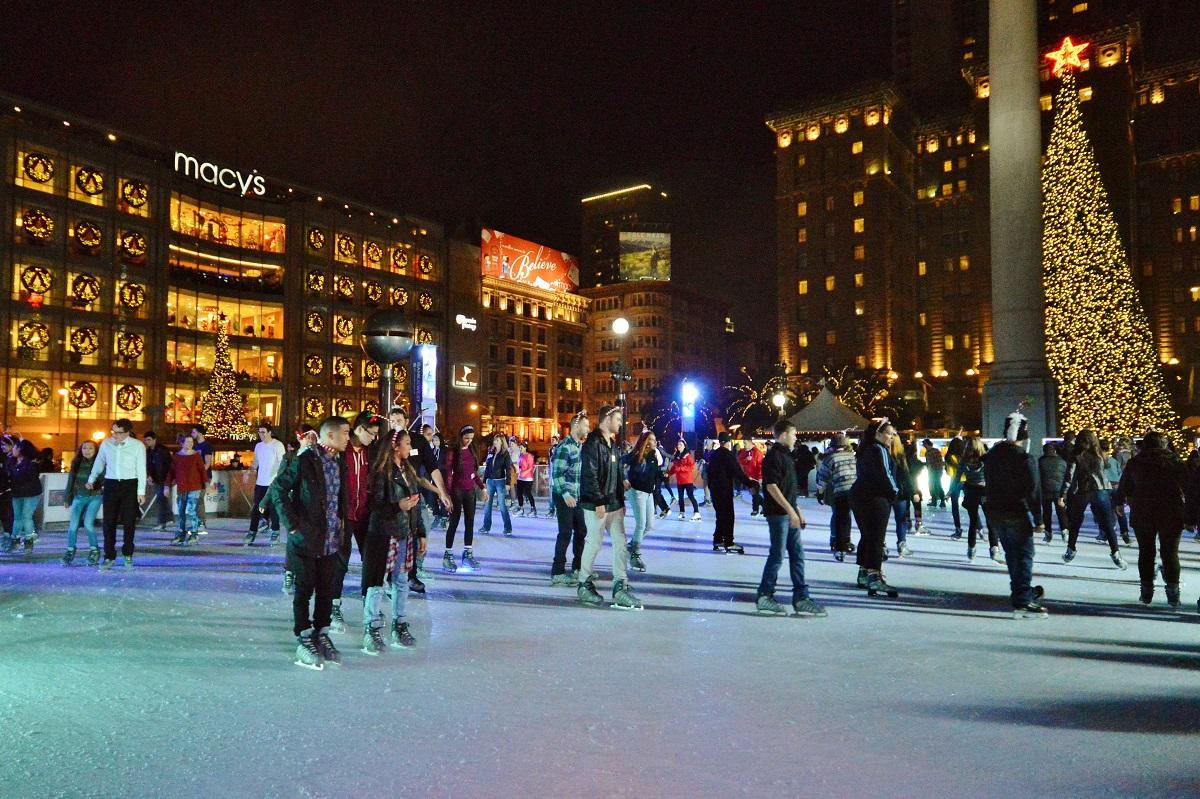 Crowds of people ice skating at an outdoor rink with Macy's building and illuminated Christmas tree visible at night.