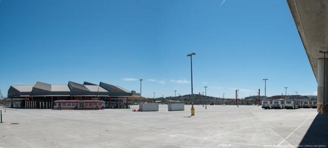 Large paved transit facility with red bus depot building, light poles, and clear blue sky