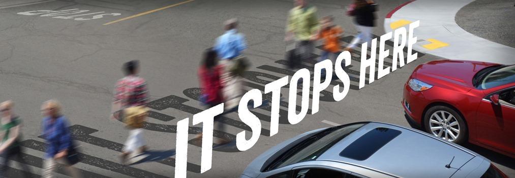 Intersection with pedestrians crossing at marked crosswalk and vehicles stopped nearby, with text "It Stops Here"
