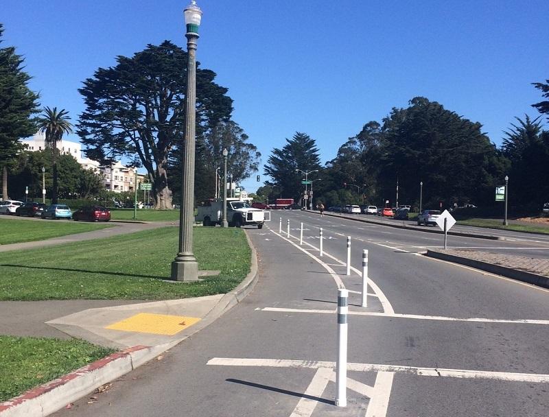 White protective posts line a bike lane on John F. Kennedy Drive with trees and park landscape visible.