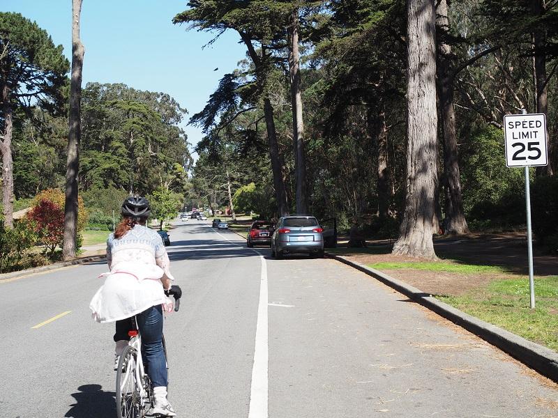 Cyclist riding on tree-lined street with 25 mph speed limit sign and parked cars visible ahead.