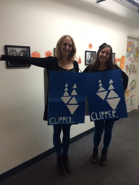 Two women hold blue Clipper card sandwich board signs in an office decorated with Halloween decorations.
