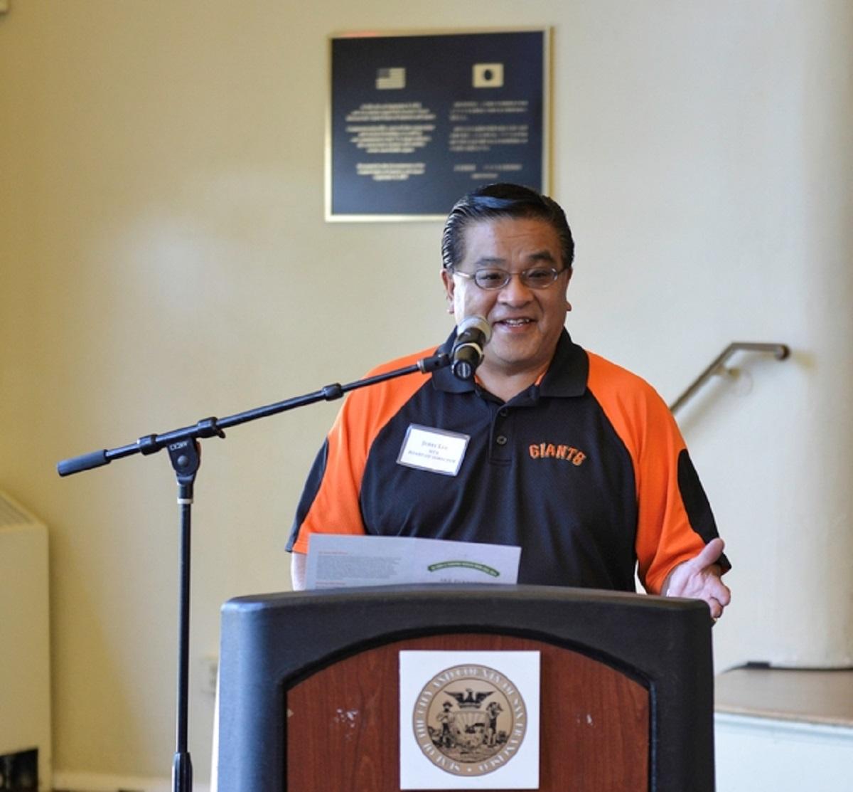 Man in SF Giants polo shirt speaking at podium with microphone at 2014 Safe Drivers Awards event.