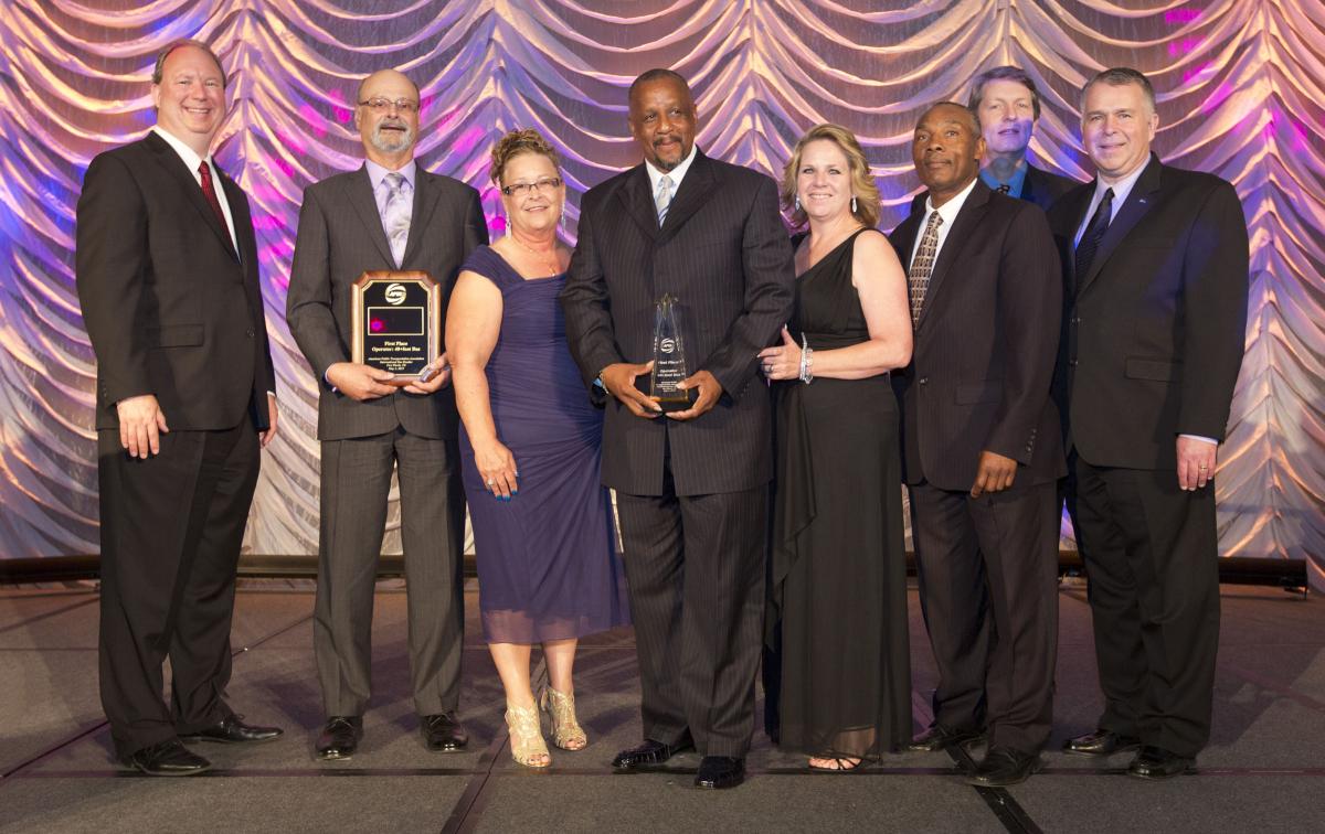 Eight people in formal attire holding awards stand in front of a purple and gold draped backdrop.