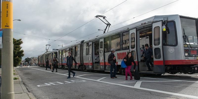 Passengers boarding and alighting from a Muni light rail train at a street-level stop with clear zone markings.