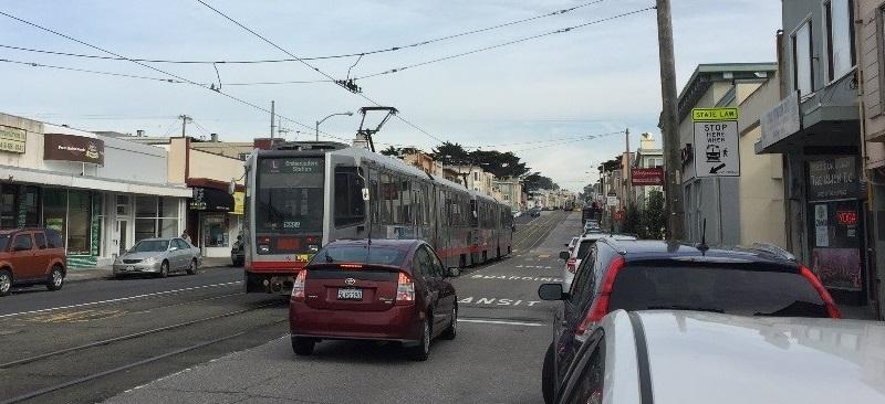 Red car stopped behind Muni light rail train at boarding zone on Taraval Street.