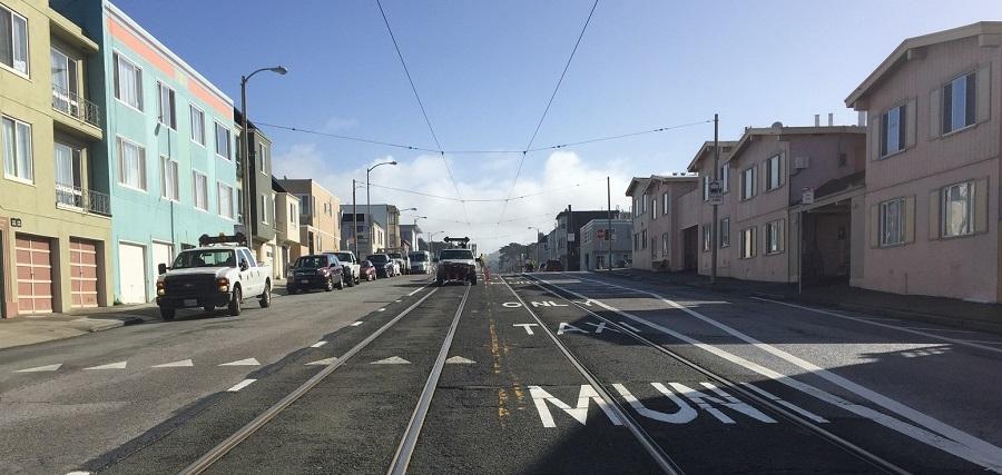 Street view of Muni light rail tracks with transit-only lane markings, parked vehicles, and residential buildings.