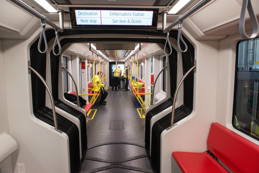 Interior of a modern light rail train car with red and yellow seats, handrails, and a digital display showing destination information.