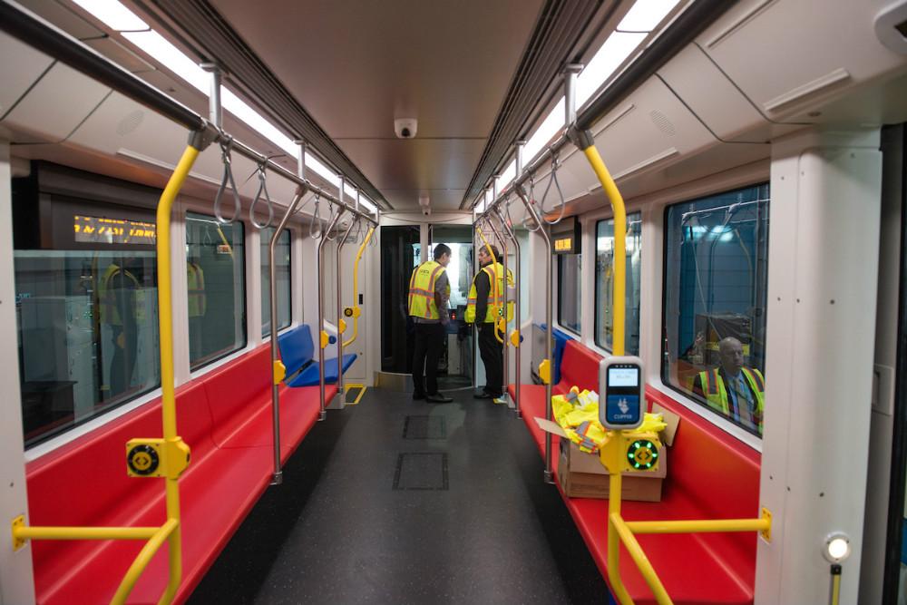 Interior of a modern red and yellow transit train car with yellow handrails, seating, and windows along both sides.