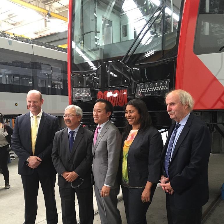 Five officials in business attire stand before a red and white light rail vehicle in a manufacturing facility.