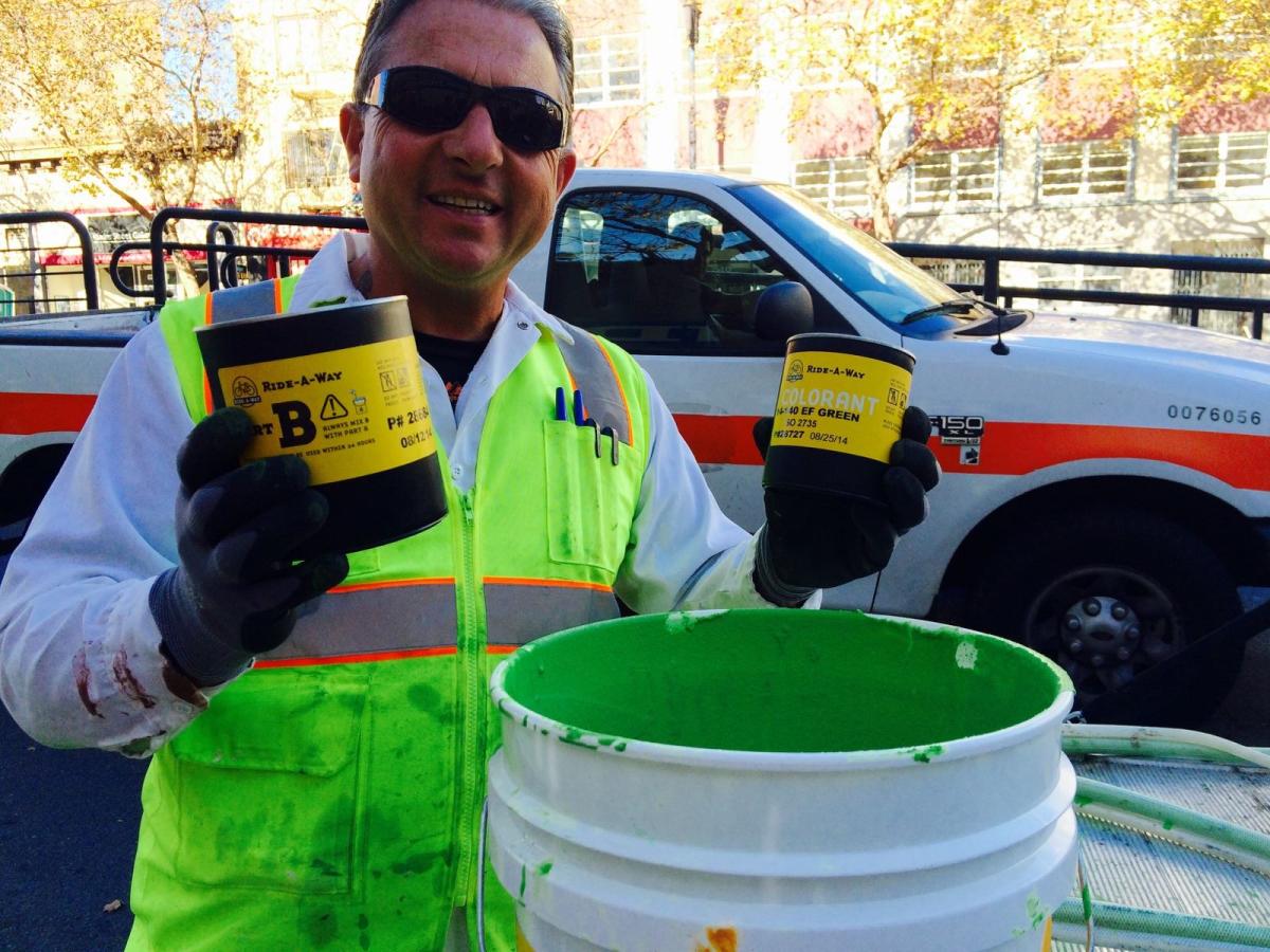 Worker in high-visibility vest holding paint cans next to a green bucket and white transit vehicle.
