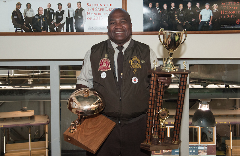 Man in transit worker uniform posing with multiple trophies and awards in front of display case.