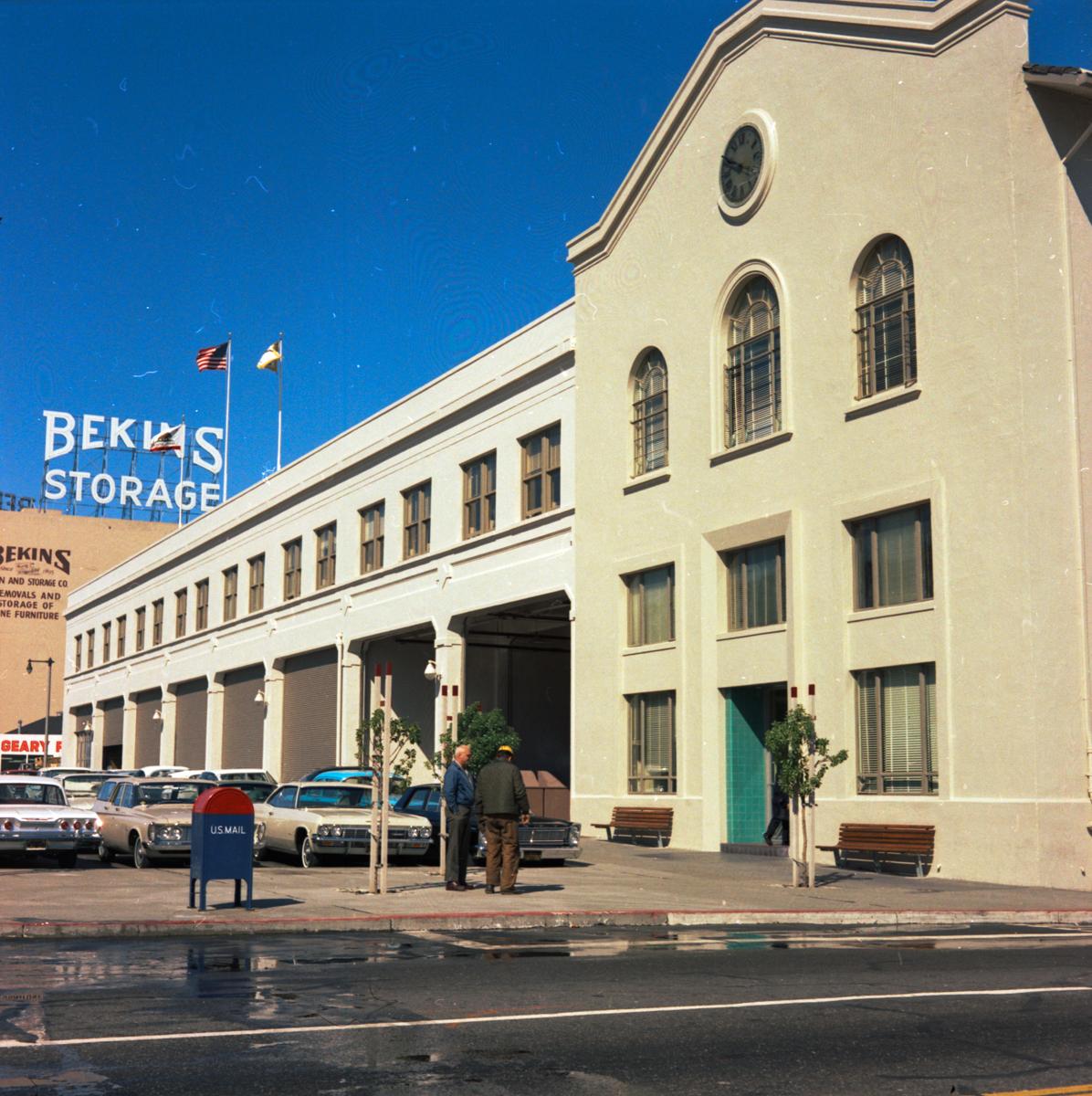 Cream-colored Presidio Carhouse building with large arched windows and garage doors, parked vintage cars, and two people on sidewalk, 1967.