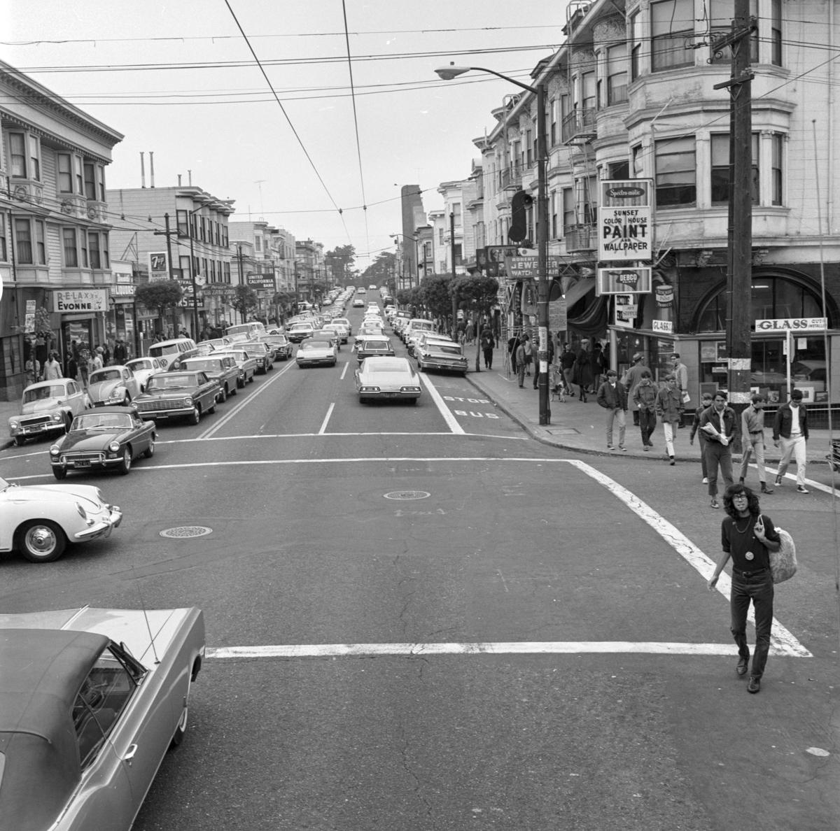 Haight and Clayton Streets intersection with pedestrians crossing, parked and moving cars, storefronts, 1967