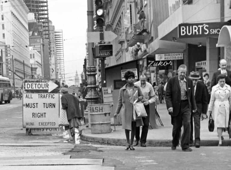 Black and white photo of pedestrians walking on Market Street during BART construction, with detour signs and storefronts visible.