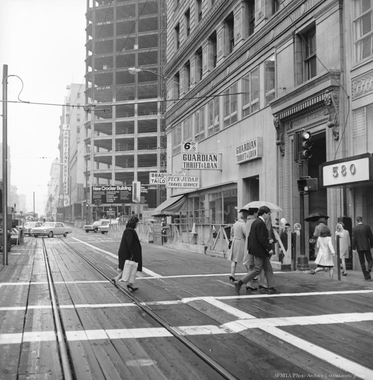 Pedestrians crossing wooden Market Street during BART construction, 1968. Streetcar tracks visible, downtown buildings in background.
