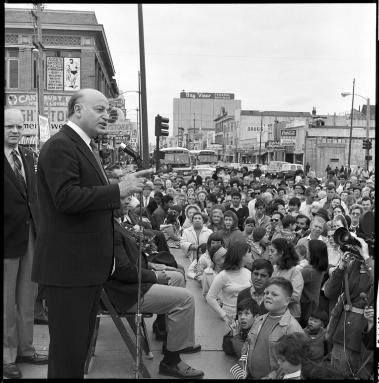 Man speaking at microphone to large seated crowd on city street with buildings and vehicles in background, 1970.